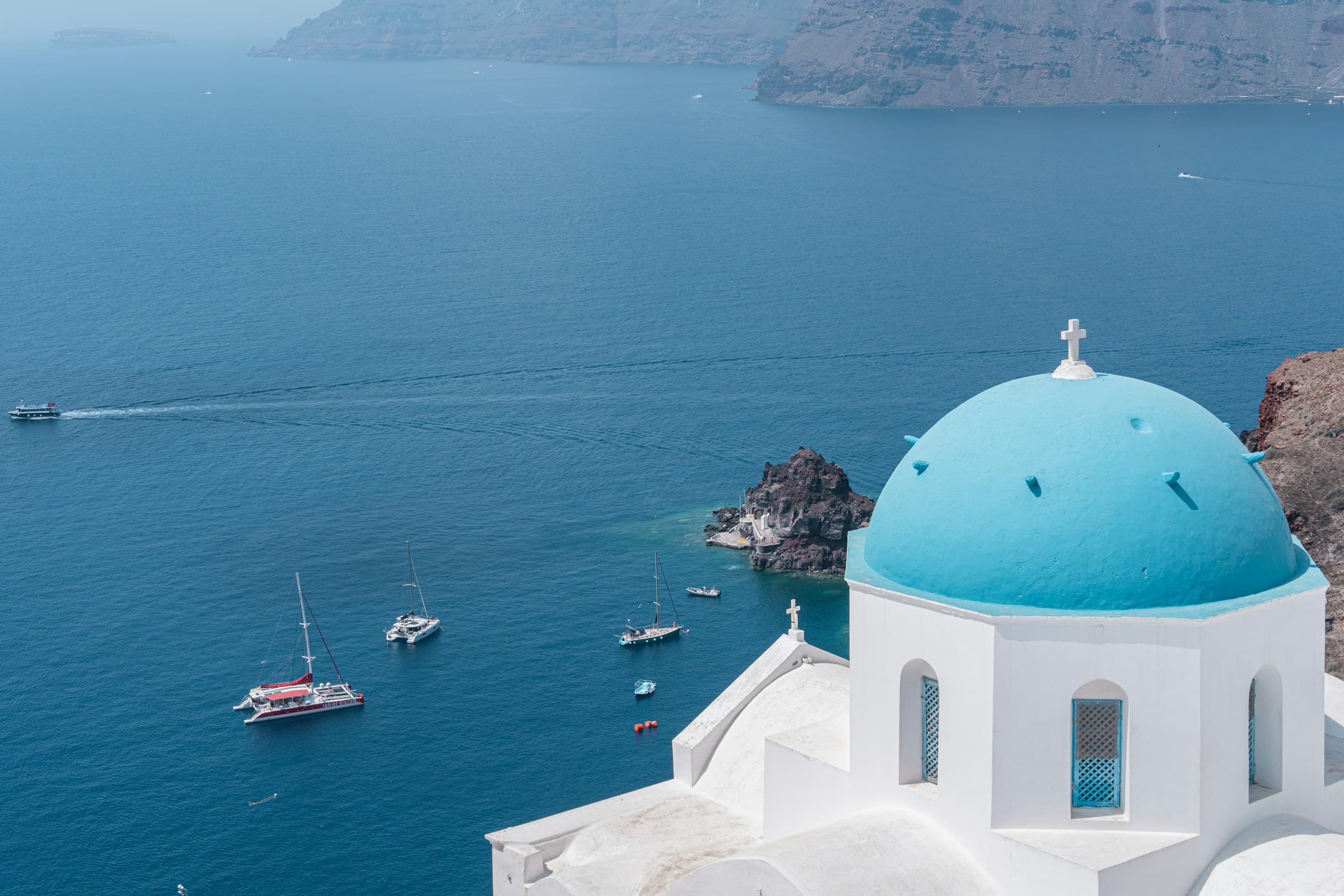 A blue-domed church above the Aegean Sea in Fira, Santorini, with sailing boats below
